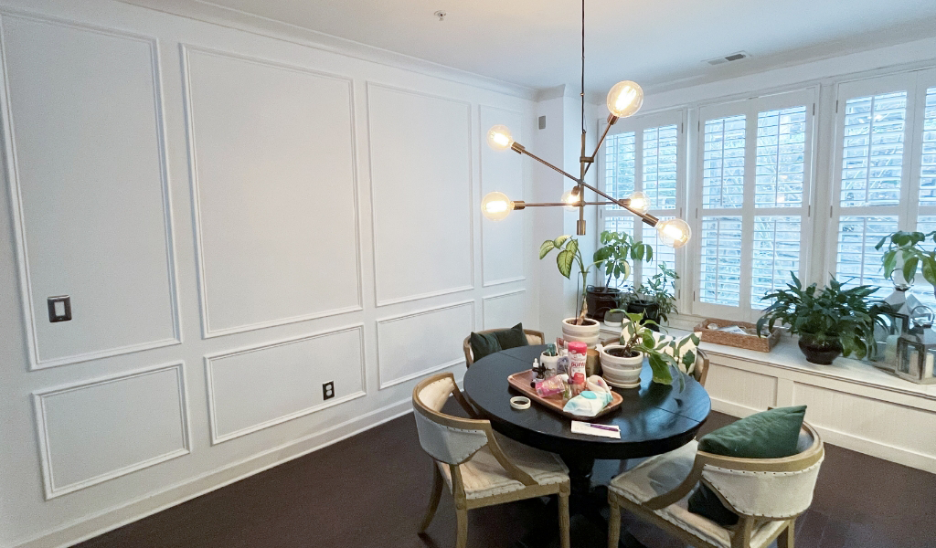 A dining room with a round black table, four chairs, and a modern chandelier is enhanced by several potted plants near large windows, offering the perfect view.