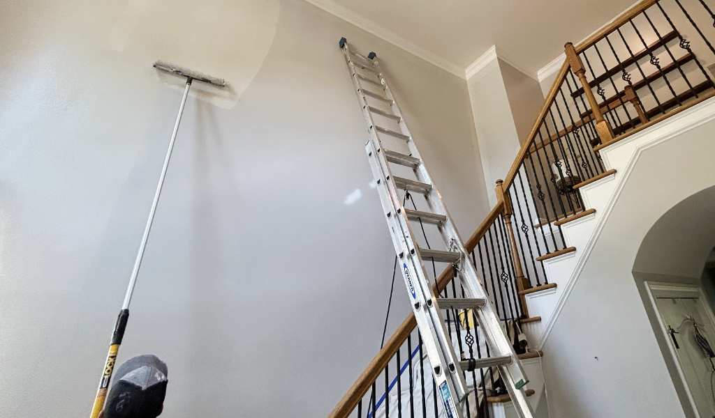 A tall ladder stands against a staircase as a worker from a painting company expertly rolls paint on a high wall with a long-handled tool, showcasing their interior painting skills.