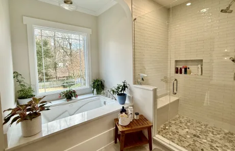 A bathroom features a large bathtub with plants nearby and a glass-enclosed shower with a tiled interior. A window provides natural light.