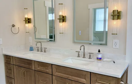 The bathroom features a double sink vanity with two rectangular mirrors and elegant brass light fixtures. The light-colored countertop adorned with soap dispensers complements the soothing beige wall.