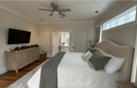 A spacious bedroom featuring a large bed with light-colored bedding, a dresser with a TV above, and a ceiling fan.