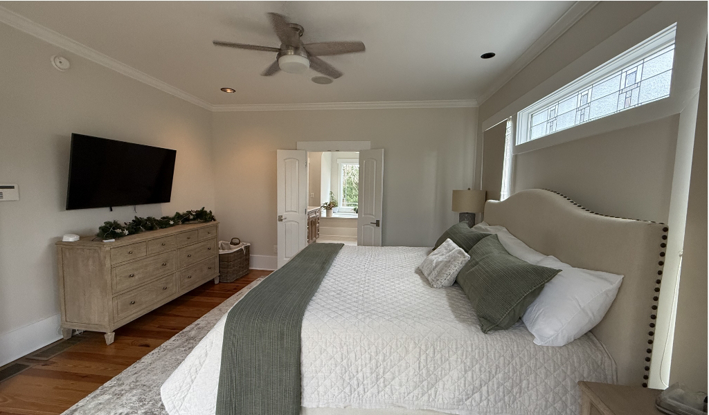 A spacious bedroom featuring a large bed with light-colored bedding, a dresser with a TV above, and a ceiling fan.