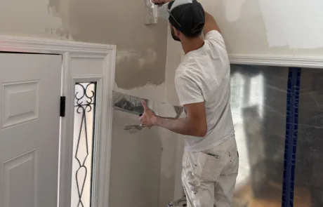 Perched on a ladder, a person applies plaster to the wall with a trowel near a decorative glass door, setting the stage for an upcoming painting project.