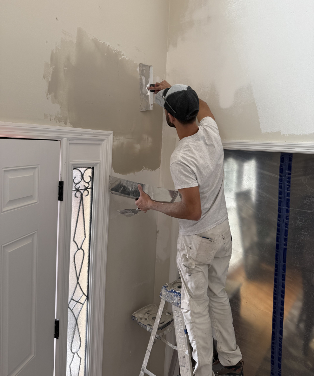 Perched on a ladder, a person applies plaster to the wall with a trowel near a decorative glass door, setting the stage for an upcoming painting project.